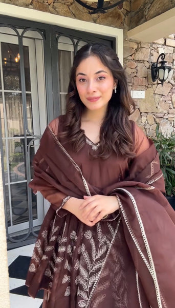 Woman wearing a brown traditional outfit with intricate patterns, standing outdoors.