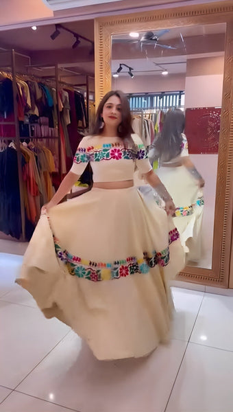 Woman in a traditional outfit with colorful top and beige skirt standing in front of a mirror.